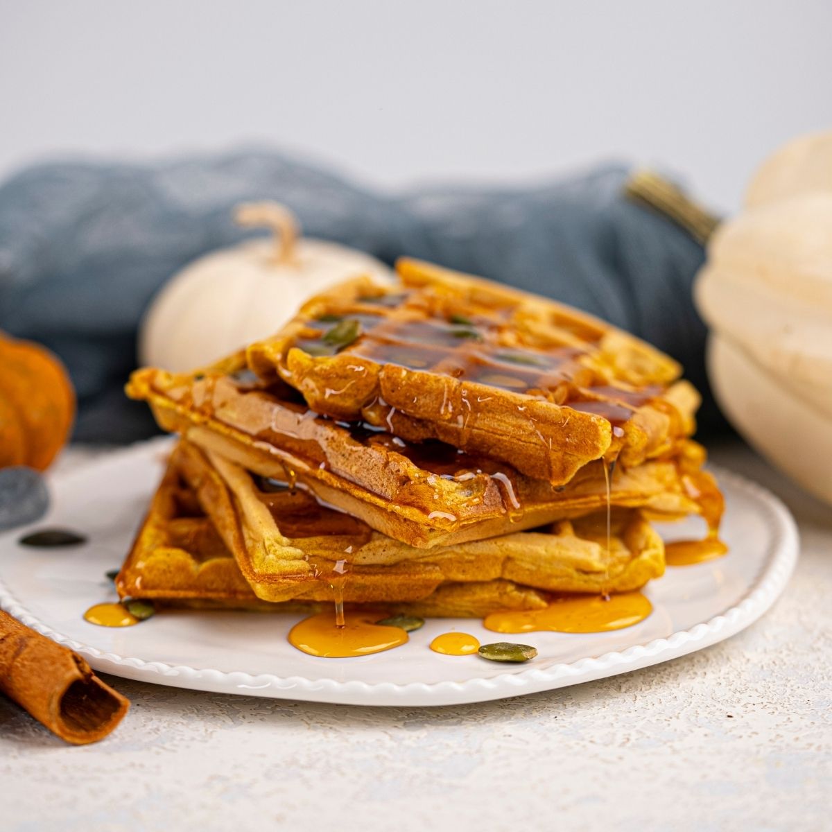 A stack of fluffy Vegan Pumpkin Waffles drizzled with syrup on a white plate, with a cinnamon stick, pumpkin seeds, and white pumpkins in the background.