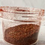 A clear plastic container partially filled with a reddish-brown Vegan Taco Seasoning, with some flavorful seasoning dusted along the inner sides and lid.