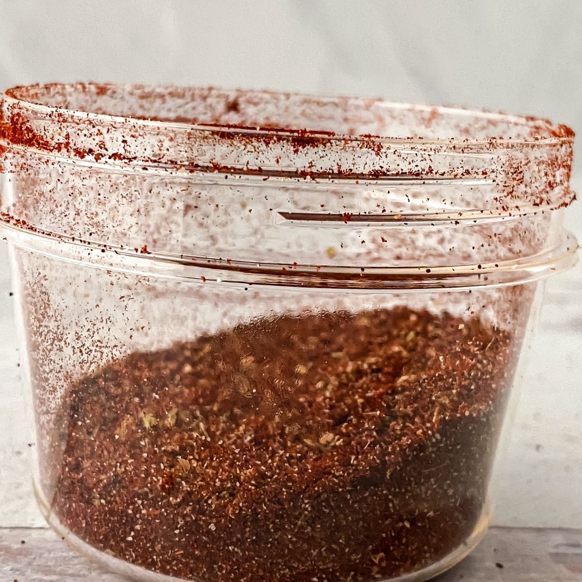 A clear plastic container partially filled with a reddish-brown Vegan Taco Seasoning, with some flavorful seasoning dusted along the inner sides and lid.