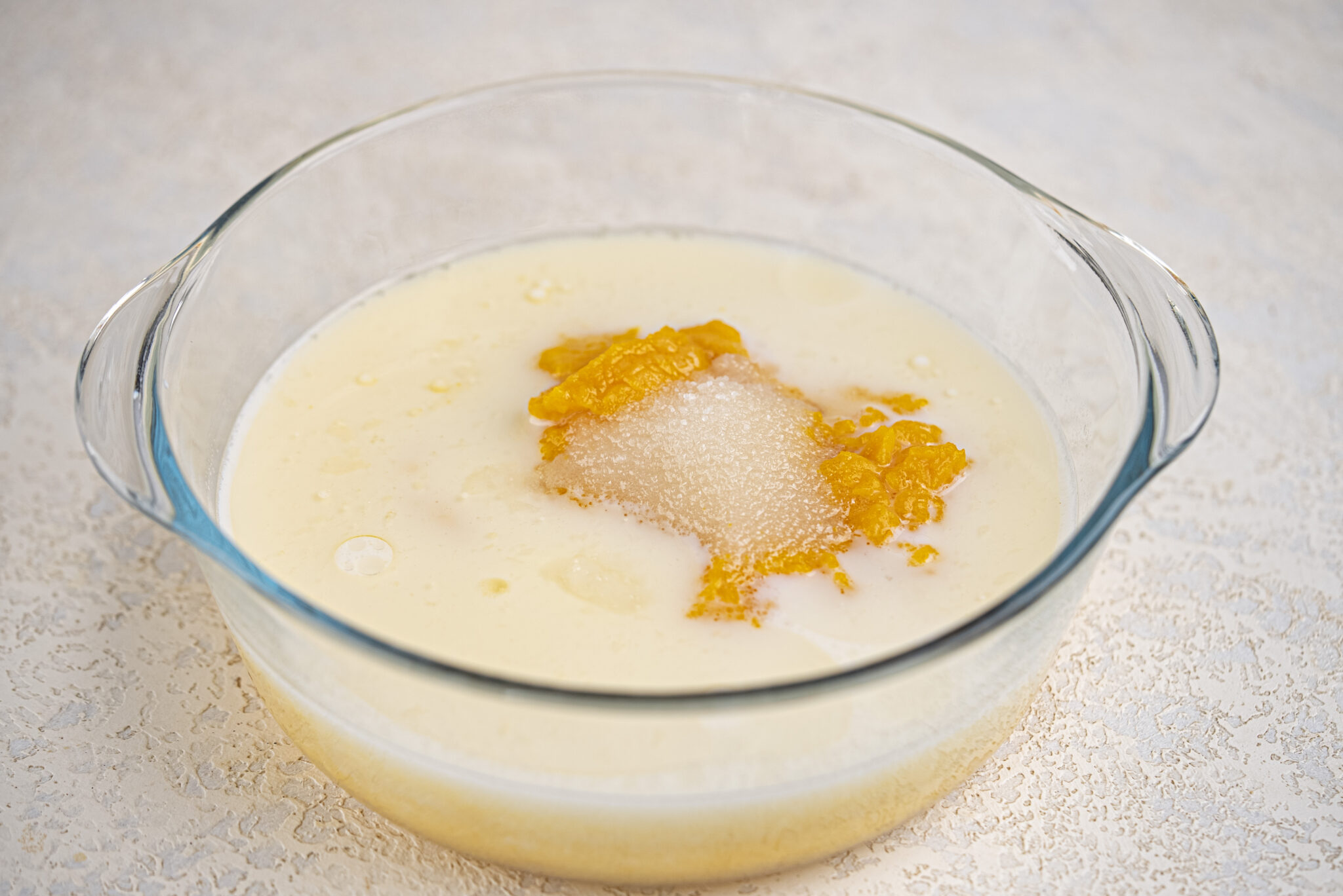 A glass bowl containing a mixture of milk, sugar, and orange-colored puree on a light textured surface.