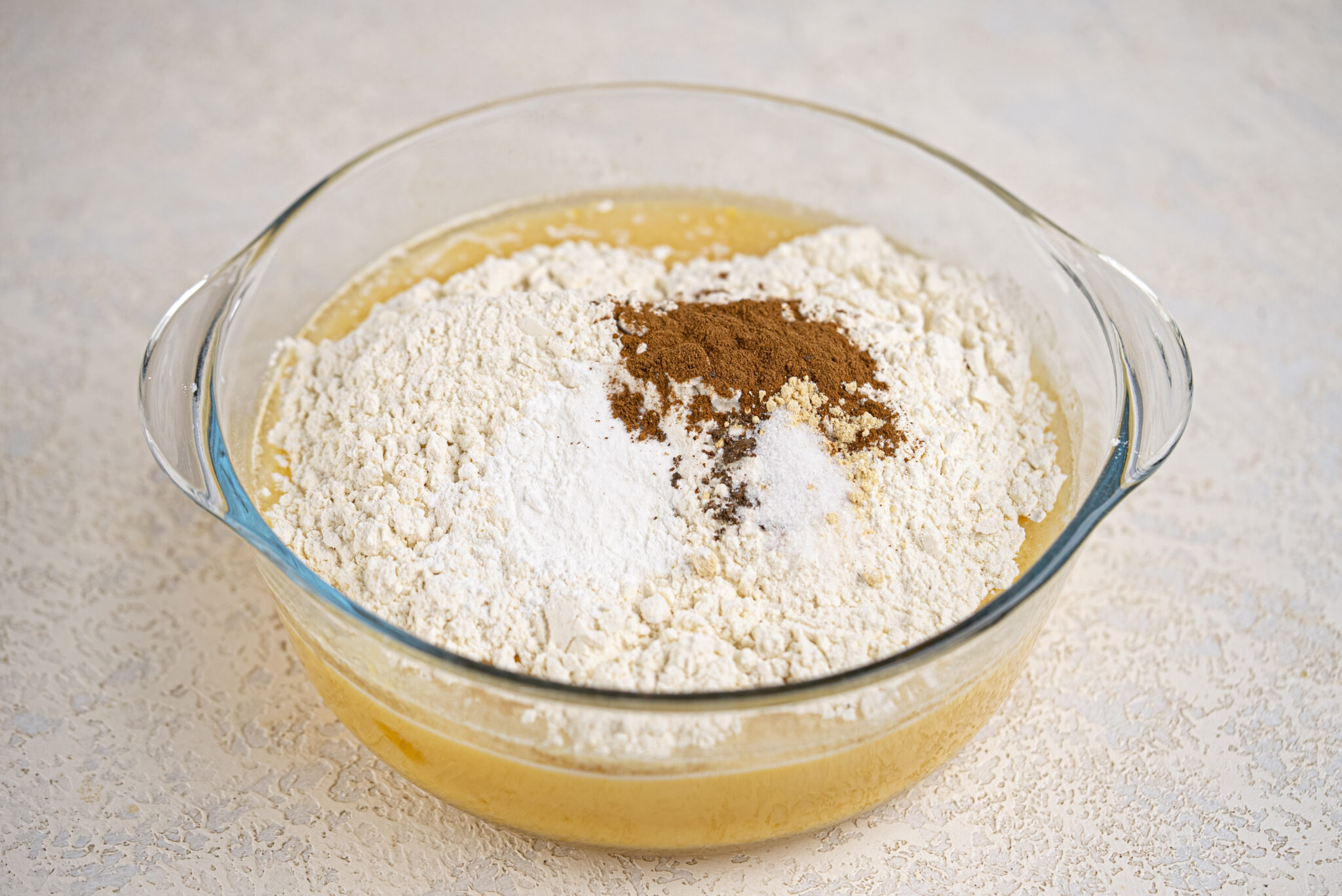 A glass bowl containing flour, baking powder, cinnamon, and salt on top of a yellow batter, ready to be mixed.