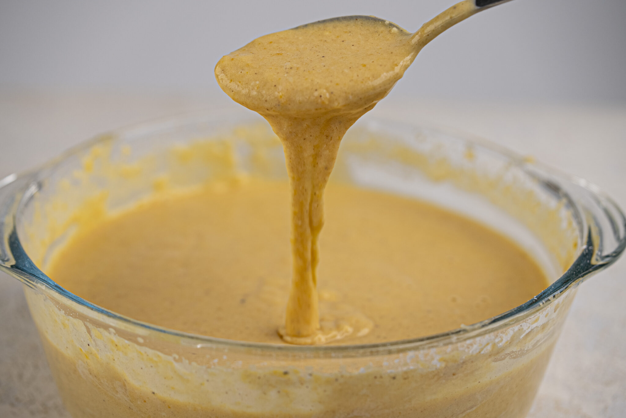 A spoon lifts thick, orange-brown batter from a glass mixing bowl, with the batter dripping back into the bowl.