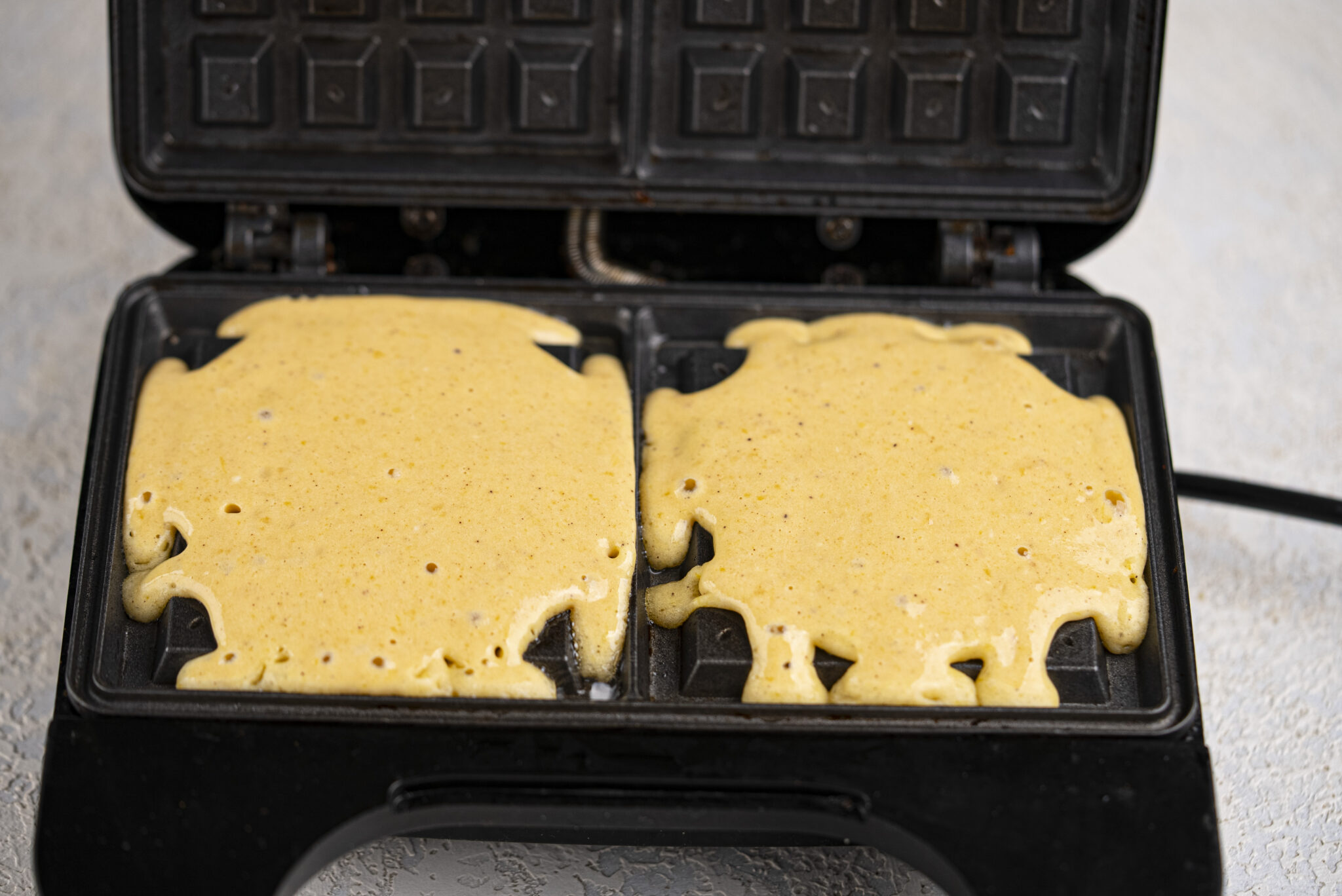 Waffle batter spread over the plates of an open waffle maker, ready to be cooked.