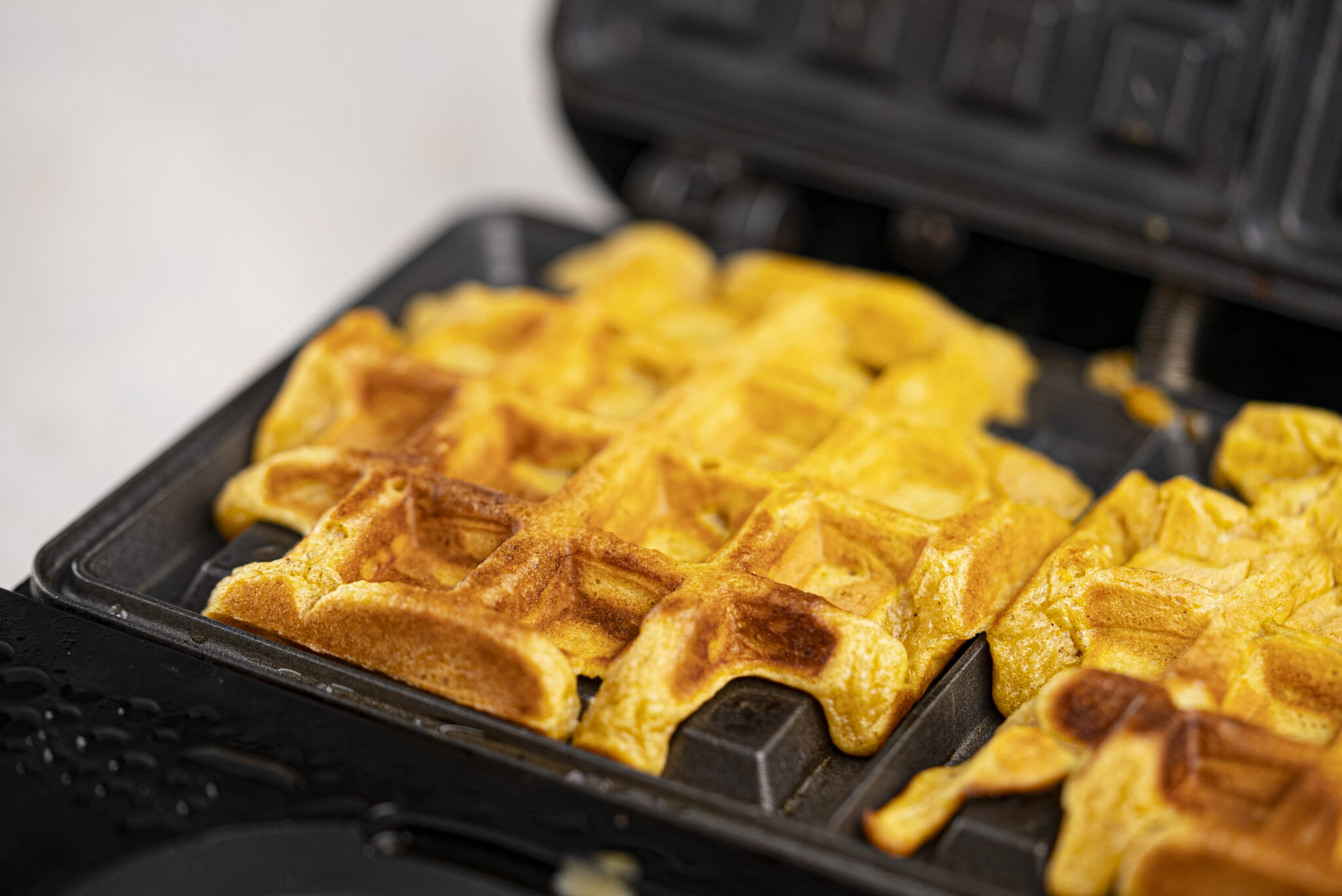 Close-up of golden waffles cooking in an open waffle iron, with some batter spilling over the edge.
