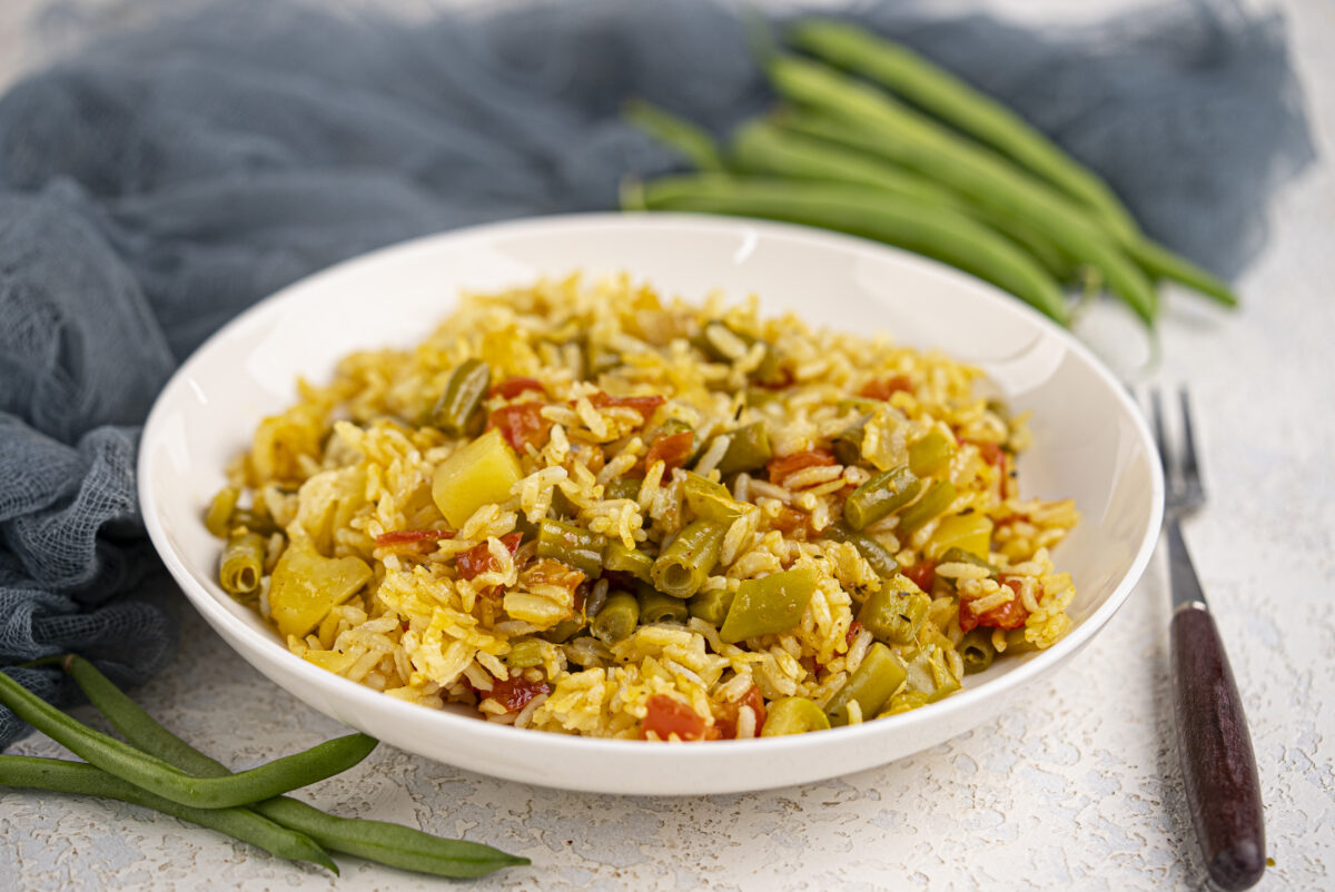 A white bowl filled with vegetable rice, including green beans, tomatoes, and yellow peppers, placed on a textured surface with raw green beans and a fork nearby.