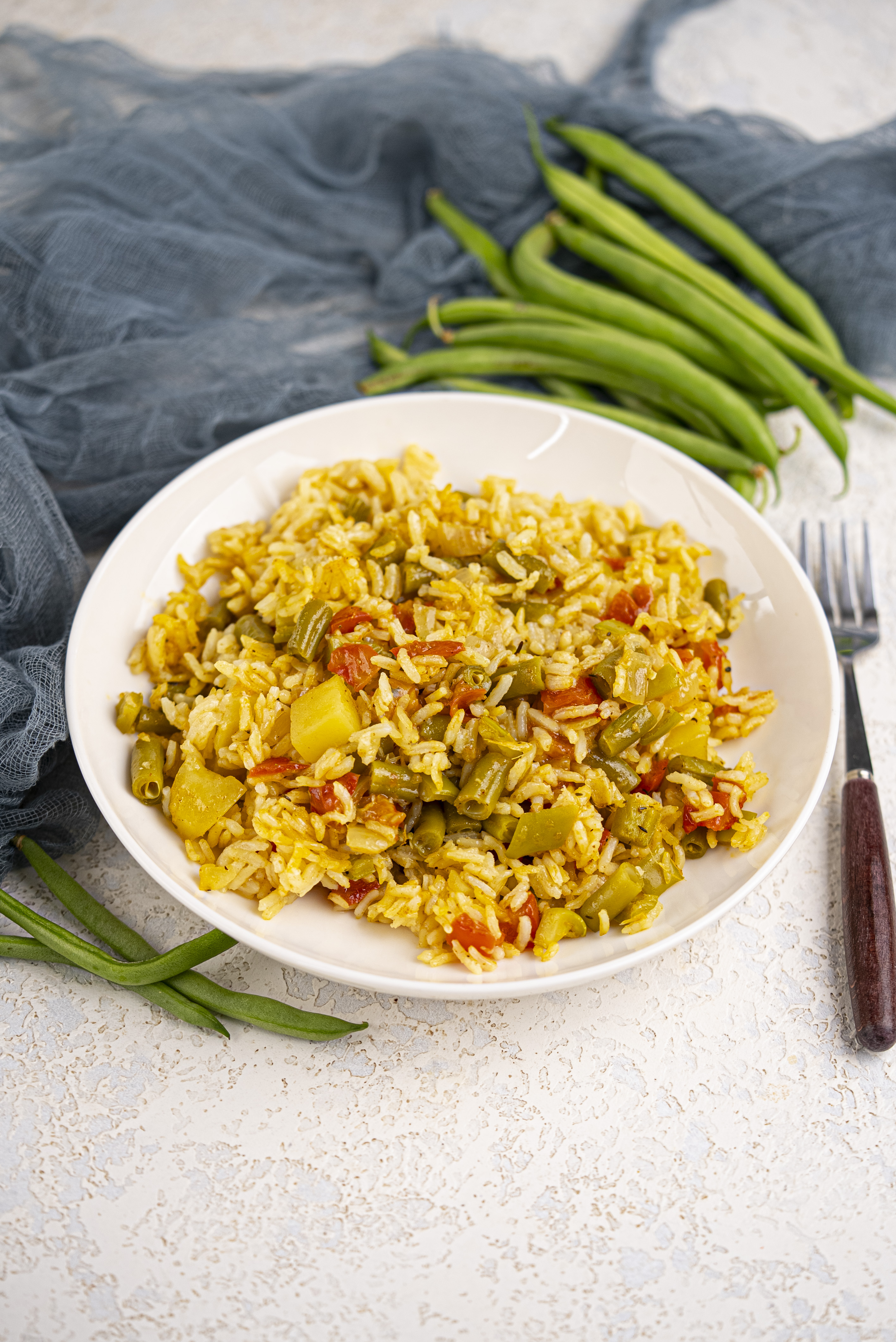 Easy Vegan Biryani Recipe | Vegetable Biryani 274 A white bowl of rice mixed with chopped green beans, tomatoes, and yellow squash sits on a light surface, with fresh green beans and a fork nearby.