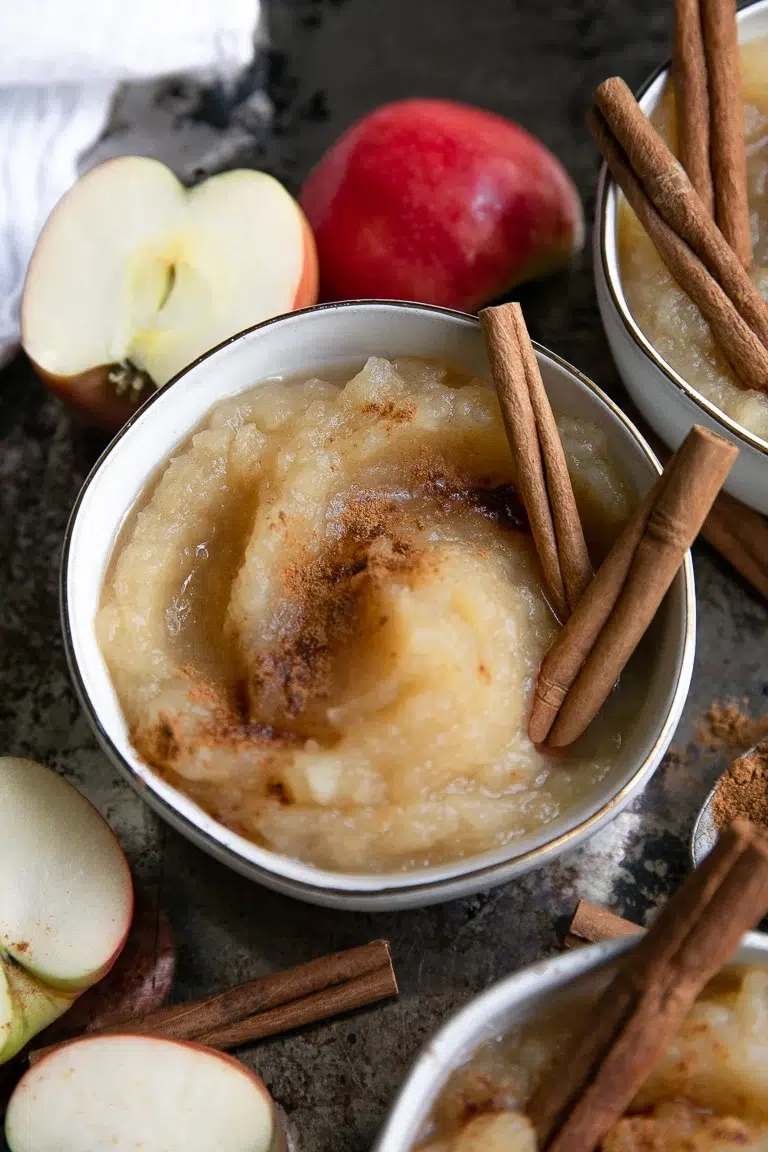 50 Delicious Vegan Desserts With Fruit That You Will Love 20 A bowl of applesauce topped with ground cinnamon and two cinnamon sticks, surrounded by fresh apple slices and a whole red apple-an inviting example of vegan desserts with fruit.