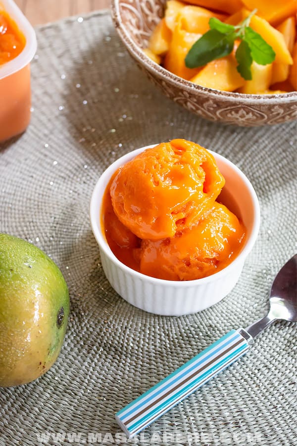 50 Delicious Vegan Desserts With Fruit That You Will Love 47 A white bowl with two scoops of orange mango ice cream, a delicious example of vegan desserts with fruit, sits on a placemat next to a mango, a spoon, and a bowl of cut fruit.