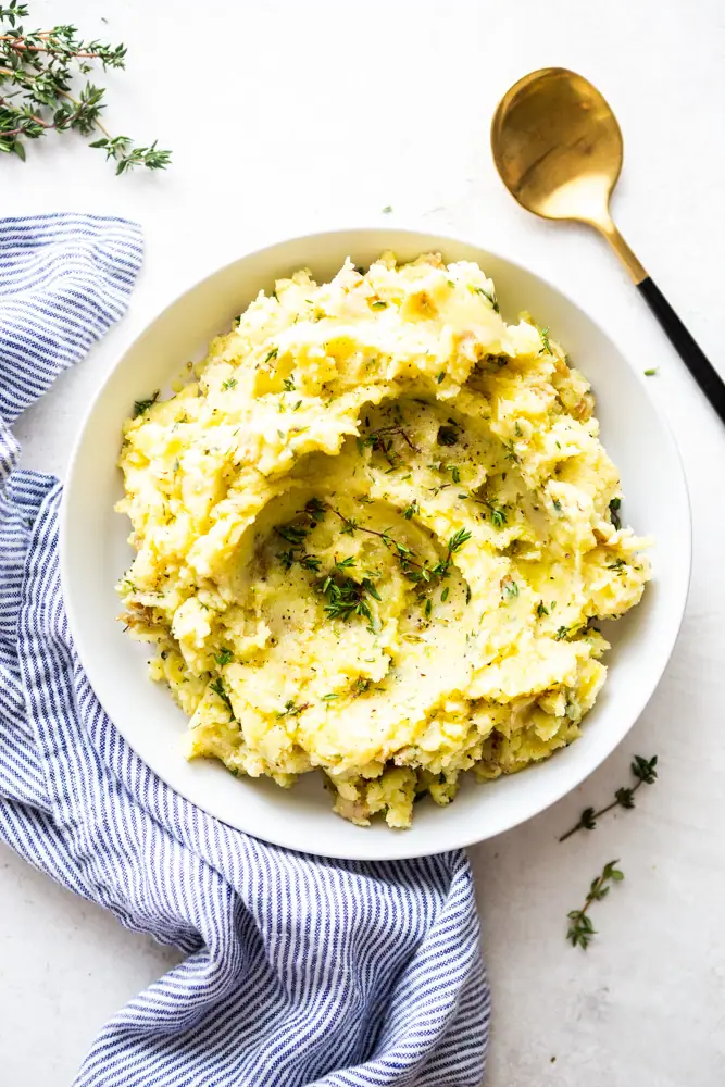 Family-Pleasing Easy Vegan Thanksgiving Recipes 14 A bowl of mashed potatoes garnished with chopped herbs, placed on a white surface with a striped towel and a gold spoon nearby.