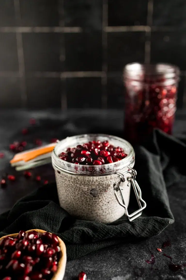 50 Delicious Vegan Desserts With Fruit That You Will Love 37 A glass jar filled with chia pudding topped with pomegranate seeds, a perfect example of vegan desserts with fruit, sits on a dark cloth, with a jar of pomegranate seeds and a spoon in the background.