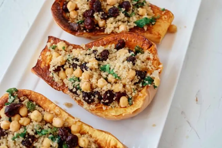 Family-Pleasing Easy Vegan Thanksgiving Recipes 26 Three roasted butternut squash halves are stuffed with a mixture of quinoa, chickpeas, raisins, and fresh herbs, served on a white rectangular plate.