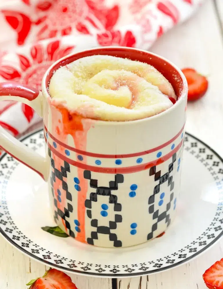 50 Delicious Vegan Desserts With Fruit That You Will Love 16 A mug cake with a swirled top sits in a patterned mug on a decorative plate, some batter spilling onto the rim. Sliced strawberries are nearby, making it a perfect choice for those who love vegan desserts with fruit.