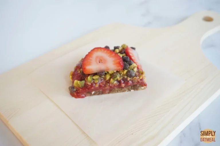 50 Delicious Vegan Desserts With Fruit That You Will Love 43 A triangular oat bar topped with sliced strawberries, chocolate chips, and chopped pistachios sits on parchment paper on a wooden cutting board-an irresistible choice for those seeking vegan desserts with fruit.