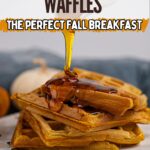 A stack of vegan pumpkin waffles is shown with syrup being poured on top, accompanied by pumpkin seeds and a blurred background featuring cozy fall decor.