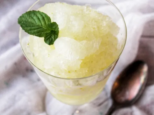 50 Delicious Vegan Desserts With Fruit That You Will Love 49 A glass of lemon granita, one of the refreshing vegan desserts with fruit, is topped with a fresh mint leaf and placed next to a metal spoon on a white cloth.