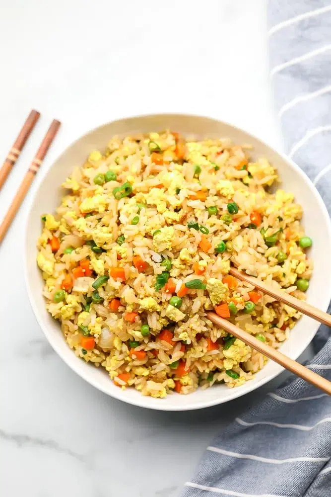 Lazy Vegan Dinner Ideas and Recipes 5 A bowl of vegetable fried rice with peas, carrots, eggs, and green onions sits beside a pair of chopsticks on a white surface-perfect inspiration for easy Vegan Dinner Ideas.