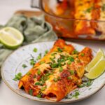 Sweet Potato and Bean Enchiladas Recipe (Vegan) 133 A plate of cheesy sweet potato and bean enchiladas topped with chopped green onions and cilantro, garnished with lime wedges, with more enchiladas in a baking dish in the background.