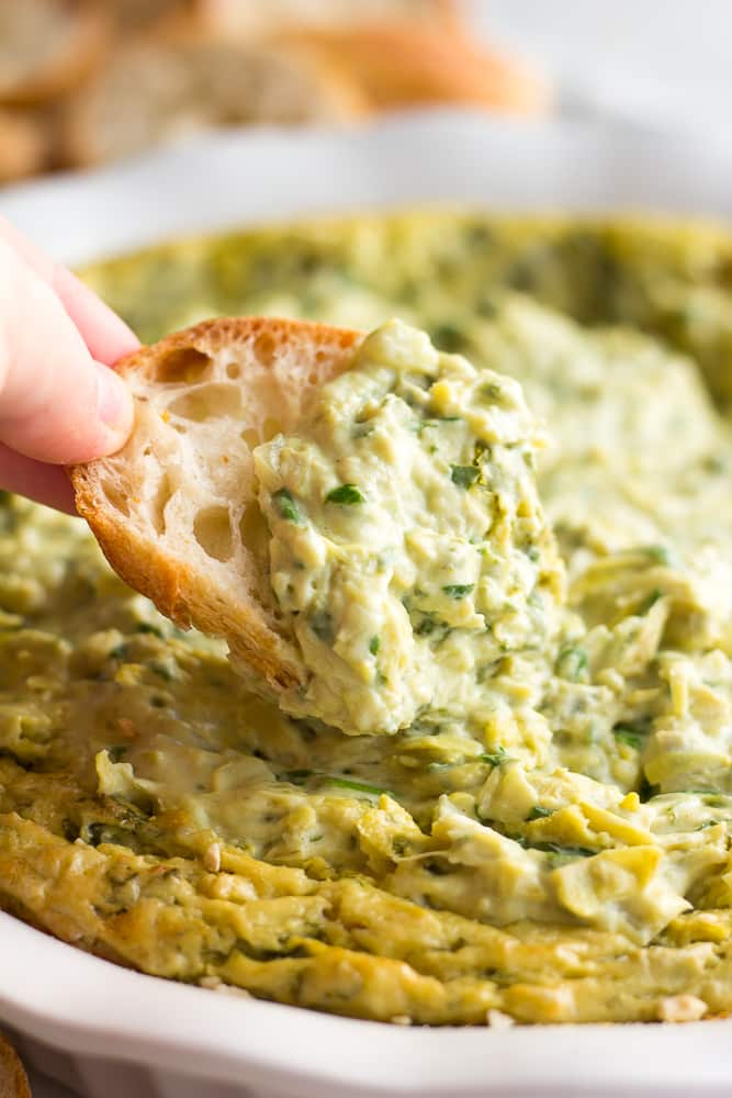 20 of The Best Easy Vegan Appetizers - Options for Everyone 13 A hand dips a slice of bread into a creamy spinach and artichoke dip in a white dish, making it one of the best vegan appetizers for any gathering.