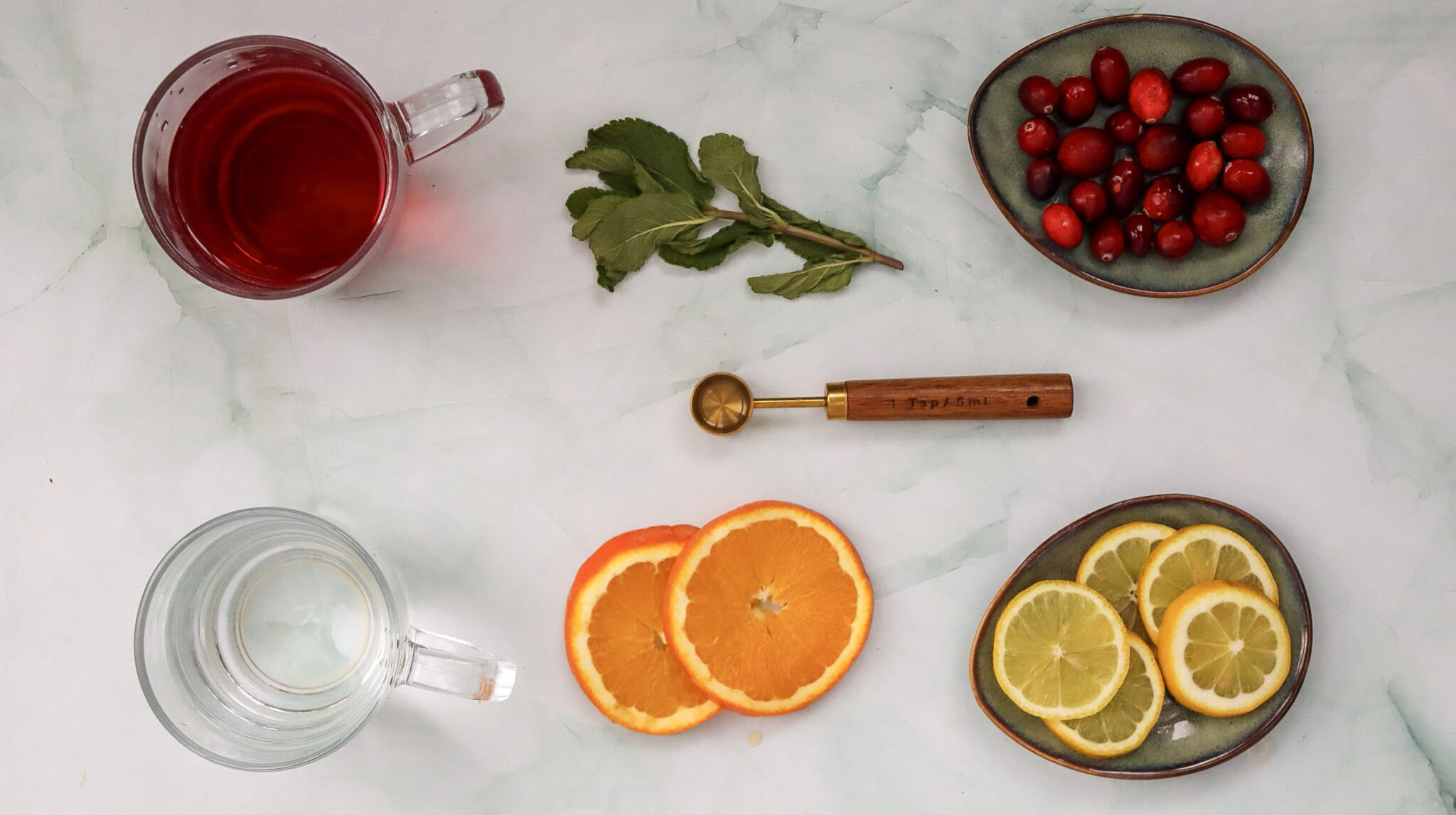 Festive Cranberry Spritzer (Spritz) Mocktail Recipe 131 Two glass cups with clear and red liquid, sliced oranges and lemons, a wooden tool, fresh mint, and cranberries arranged neatly on a light surface.