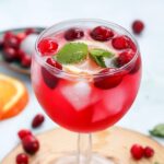 A glass of cranberry spritzer with ice cubes, cranberries, orange slice, and mint leaves sits on a wooden surface, surrounded by fresh cranberries and a slice of orange in the background.