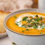 A bowl of creamy vegan roasted pumpkin soup, garnished with pumpkin seeds, chopped herbs, and a swirl of cream.