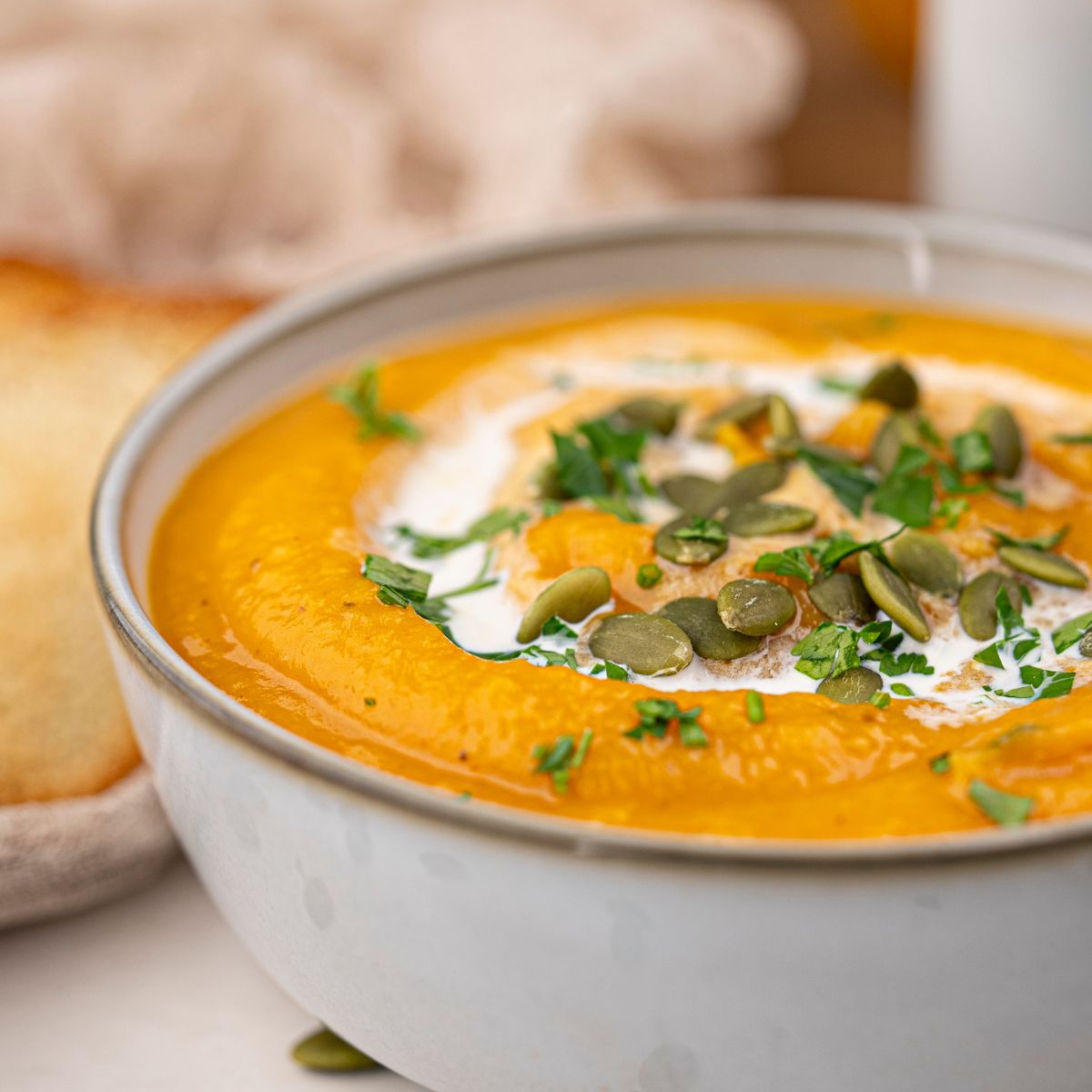 A bowl of creamy vegan roasted pumpkin soup, garnished with pumpkin seeds, chopped herbs, and a swirl of cream.