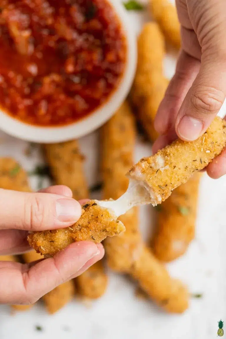 20 of The Best Easy Vegan Appetizers - Options for Everyone 17 A hand pulls apart a vegan mozzarella stick with gooey cheese stretching inside, perfect for Easy Vegan Appetizers. A bowl of marinara sauce and more vegan mozzarella sticks sit in the background.