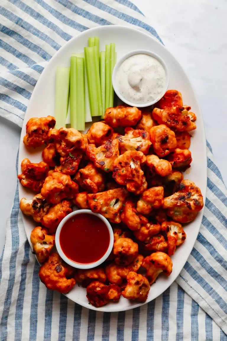 20 of The Best Easy Vegan Appetizers - Options for Everyone 19 A white oval plate with buffalo cauliflower bites, celery sticks, ranch dip, and a bowl of red dipping sauce sits on a blue-and-white striped cloth-one of the best vegan appetizers for any gathering and perfect for fans of easy vegan recipes.