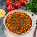 A bowl of authentic chickpea stew garnished with chopped herbs, surrounded by fresh tomatoes, parsley, and a halved lemon on a light surface—perfect for fans of vegan chana masala.