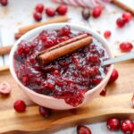 A bowl of cranberry sauce garnished with a cinnamon stick sits on a wooden board, surrounded by whole cranberries and cinnamon sticks.