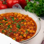 A bowl of Vegan Chana Masala garnished with fresh coriander, with tomatoes and herbs in the background. Text reads: “Chana Masala - Quick, Easy & Authentic Chickpeas.”.