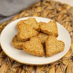 A white plate holds several heart-shaped Gluten Free Valentine's Biscuits topped with sesame seeds, arranged on a woven placemat.