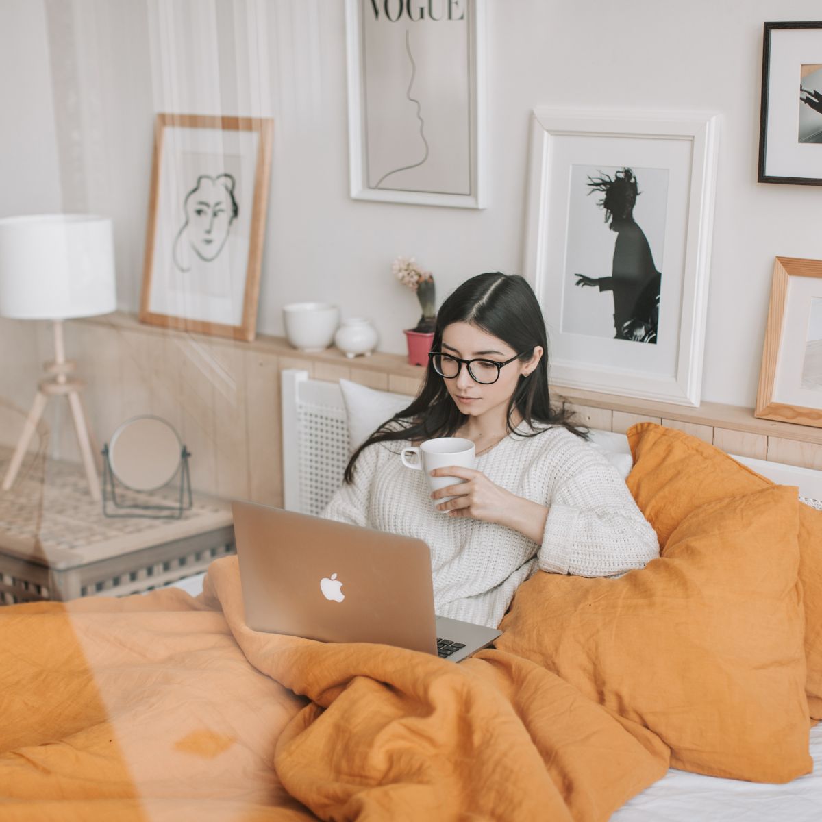 A woman sits in bed with an orange blanket, working on a laptop and drinking from a white mug, enjoying her daily routines at home in a room decorated with framed art.