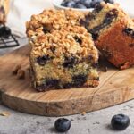 A close-up of two Vegan Blueberry Coffee Cake slices on a wooden board, with fresh blueberries scattered nearby.