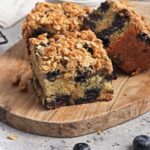 Two pieces of vegan blueberry coffee cake with crumb topping are displayed on a wooden board, blueberries scattered nearby. Text above says "Blueberry Coffee Cake" and "Vegan Recipe".