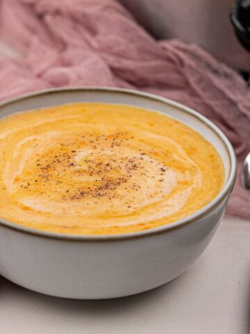 A bowl of creamy orange vegan soup topped with a sprinkle of black pepper, placed next to a spoon and a pink cloth.