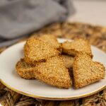 A plate of heart-shaped, sesame-coated keto biscuits on a woven placemat, with text reading "Keto Valentine's Biscuits" and "Vegan gluten free Valentine's biscuits.