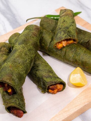 Four green rolled flatbreads filled with a cooked mixture, served on a wooden board with lemon wedges and a green chilli pepper—perfect for anyone craving an easy taquitos recipe.
