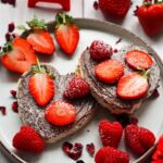 A plate with two heart-shaped pastries topped with chocolate spread and sliced strawberries, surrounded by fresh strawberries and raspberries.