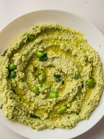 Quick And Easy Edamame Hummus Recipe 135 A white bowl filled with green houmous, garnished with whole edamame beans, chopped herbs, and a drizzle of olive oil.