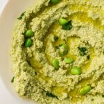 A bowl of edamame houmous topped with edamame beans, chopped herbs, and a drizzle of oil. Text at the bottom reads: "Edamame Houmous Recipe @wemadethisvegan." Try this quick recipe for a healthy snack!.