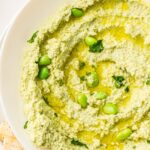 A bowl of green edamame houmous drizzled with olive oil and garnished with whole edamame, served next to crackers; text reads “Edamame Houmous Recipe – an easy recipe for a quick, tasty snack.”.