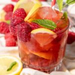 A glass of raspberry and lemon cocktail with ice, garnished with fresh raspberries, lemon slices, mint leaves, and a striped straw.