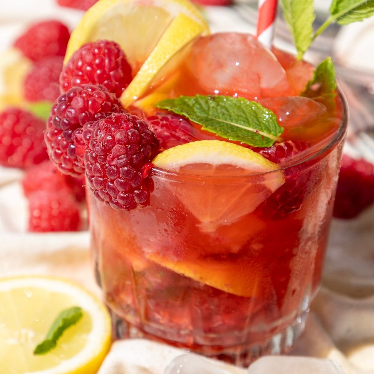 A glass of raspberry mojito with ice, garnished with fresh raspberries, lemon slices, mint leaves, and a striped straw.