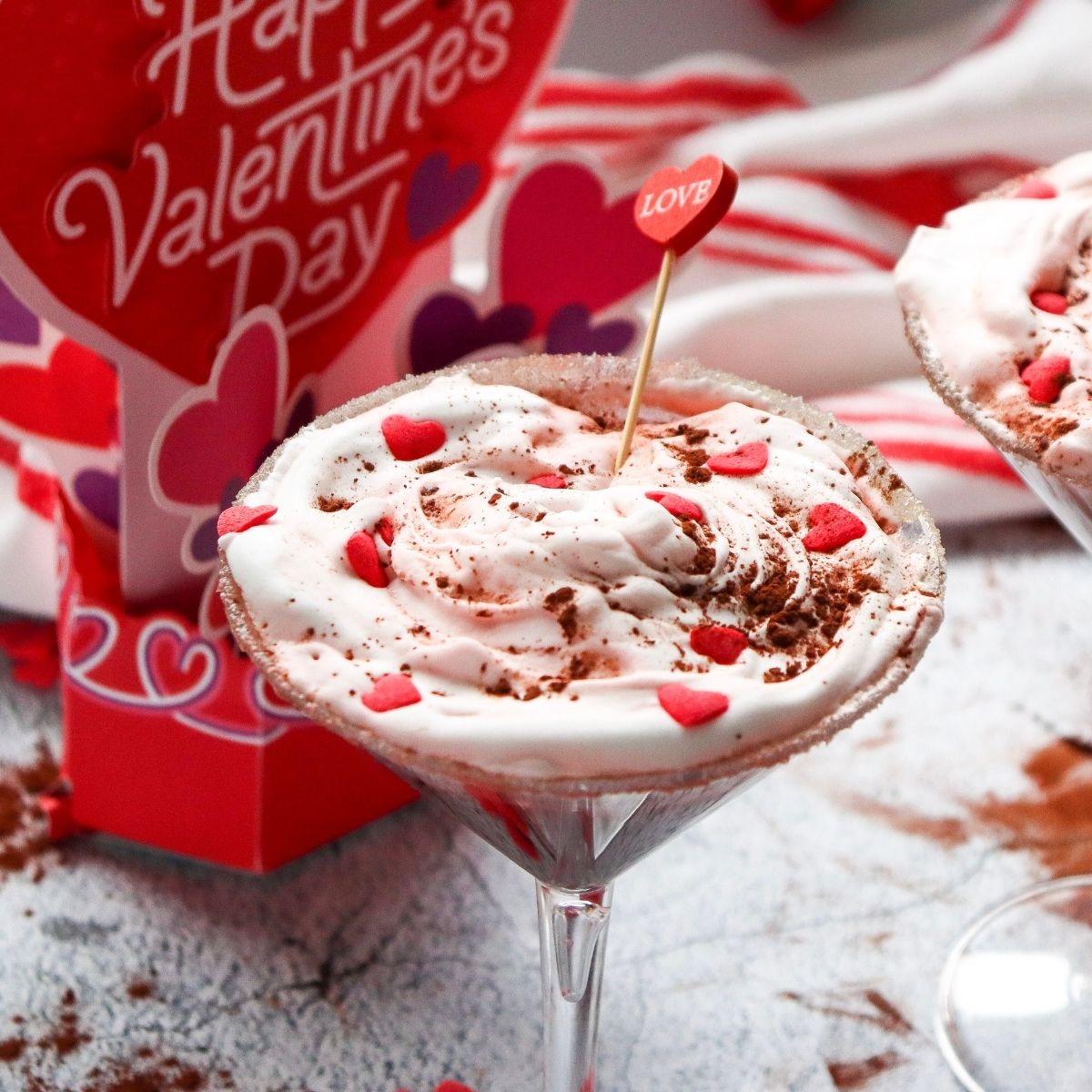 red velvet martinis topped with pink heart sprinkles, cocoa powder, and a cocktail stick with a “LOVE” heart; a Valentine’s Day card is in the background.