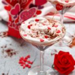 A martini glass filled with a creamy Red Velvet Martini Mocktail, topped with whipped cream and heart-shaped sprinkles, sits beside a red rose and festive decorations—a delightful mocktail recipe for any celebration.