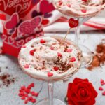 Two martini glasses filled with Red Velvet Martinis topped with whipped cream and heart sprinkles, set on a Valentine’s Day-themed table alongside a rose and a card—perfect for a romantic toast.