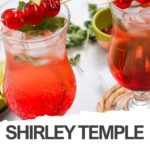 Two Shirley Temple mocktails in curved glasses garnished with cherries, mint leaves, and lime slices, set on a white table—an ideal Valentine’s drink with a fresh twist. A green bowl and herbs provide the perfect backdrop for this classic mocktail recipe.