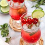 Two glasses filled with ice and vibrant red mocktail—perfect as a Valentine’s Drink—are garnished with lime slices, mint leaves, and skewered cherries, set on a table with extra limes and cherries nearby.