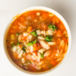 A bowl of Vegan Minestrone Soup with vegetables, beans, and pasta in a tomato broth, seen from above. Text reads "Vegan Minestrone Soup." Perfect for making in your slow cooker.